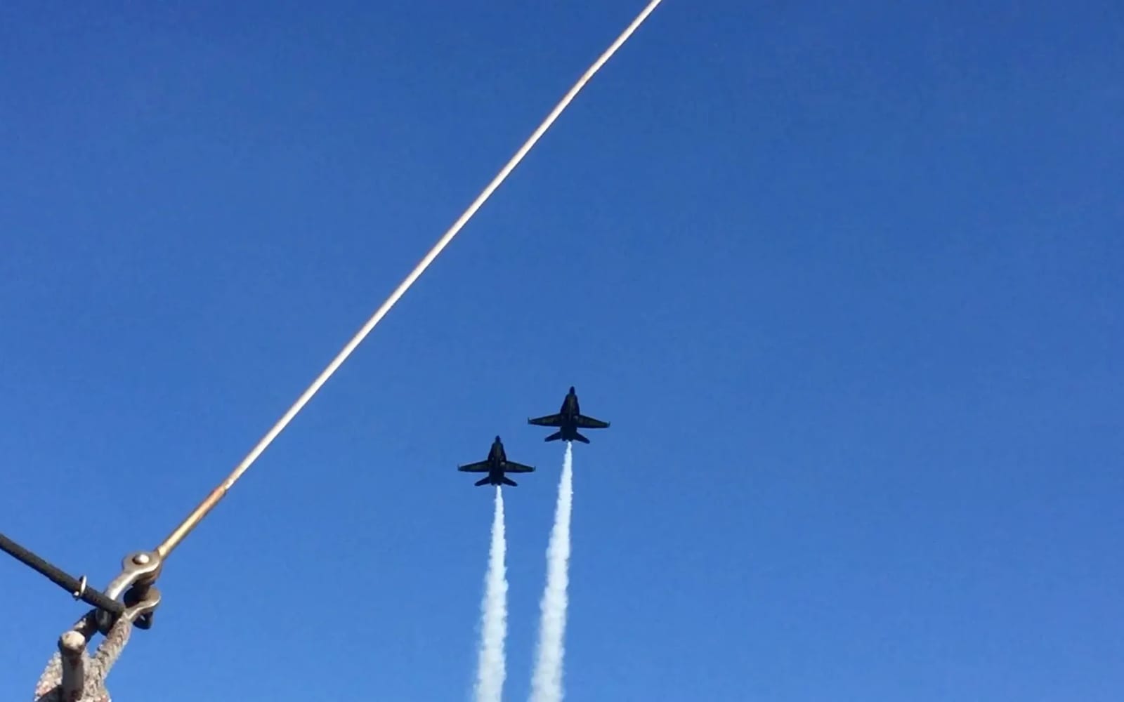Two Blue Angels jets flying past with smoke trails, seen from the deck of a sailboat past the rigging